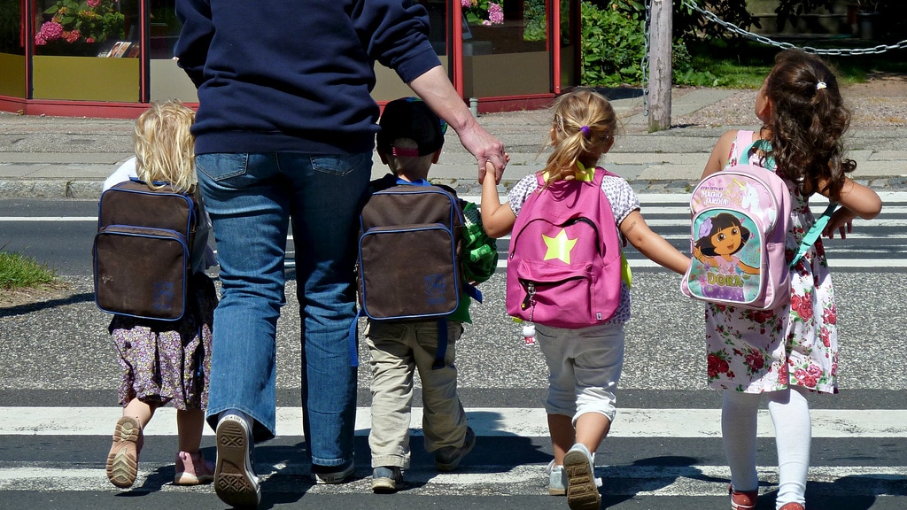 Four children wearing backpacks walking in a cross walk all holding hands with each other and an adult