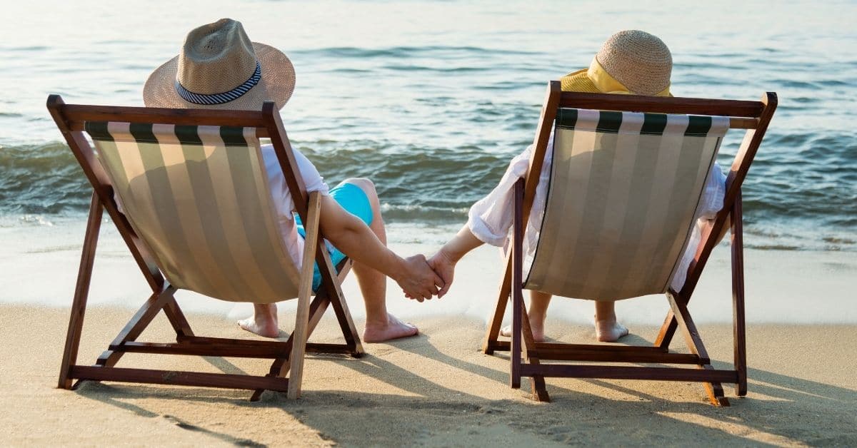 Couple holding hands sitting in folding chairs wearing hats watching sunset on the beach along shoreline loving their location
