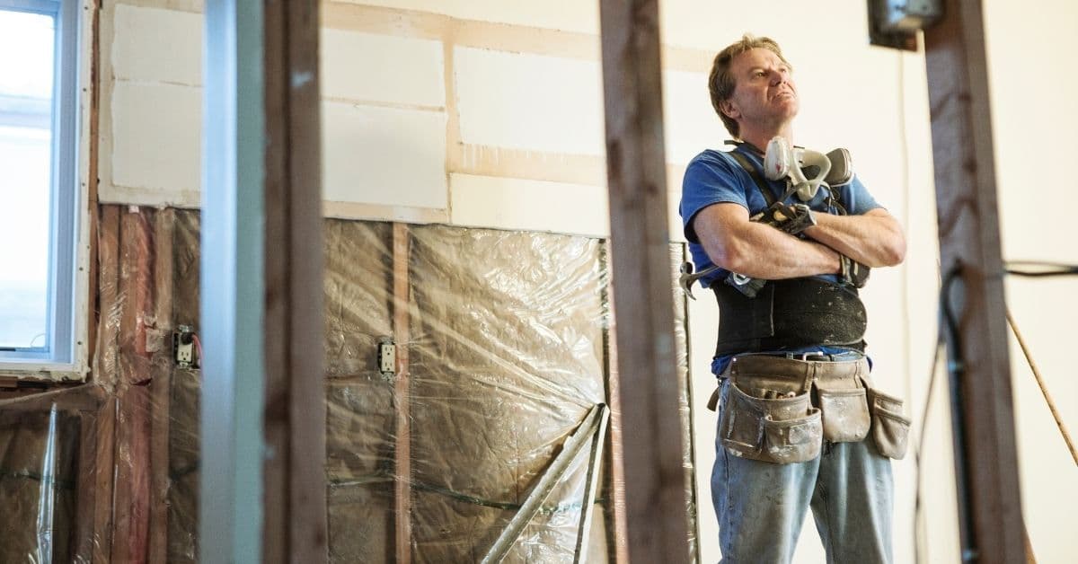 man wearing tool belt arms folded inspecting home renovation project inside house