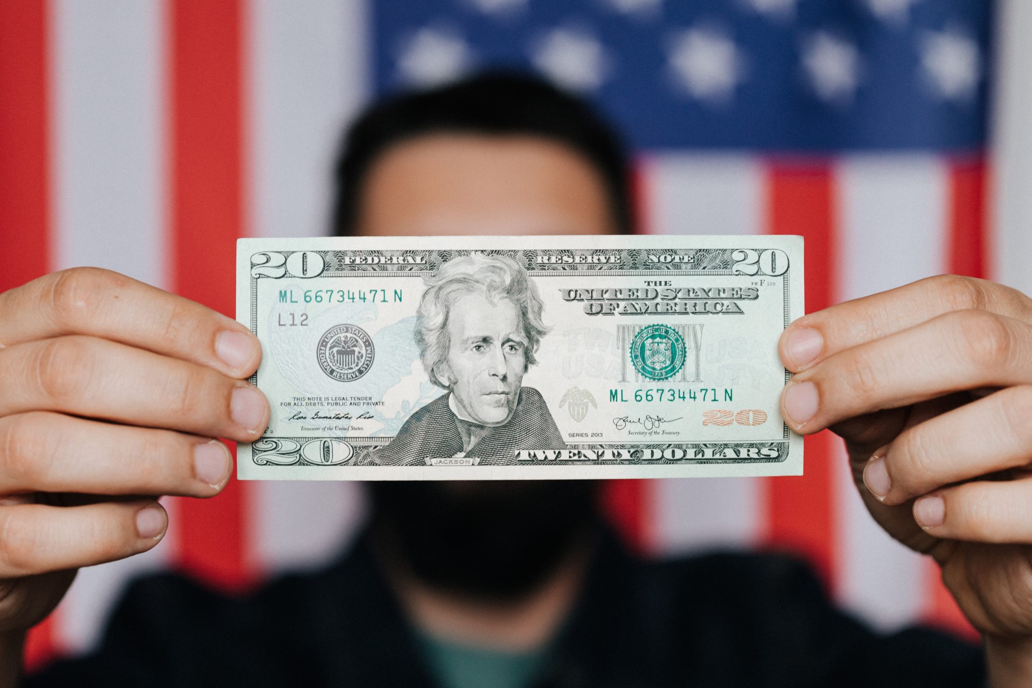 A man standing in front of an American flag holding a twenty dollar bill in front of his face