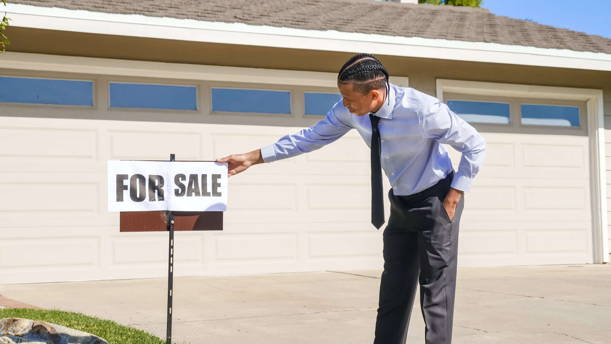 A black male real estate agent stands outside a home leaning over to look at a For Sale sign sticking out of the ground.