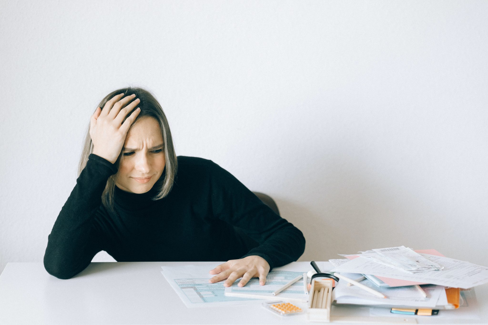 a woman sits at a desk with paperwork with her hand on her head and a scowling expression