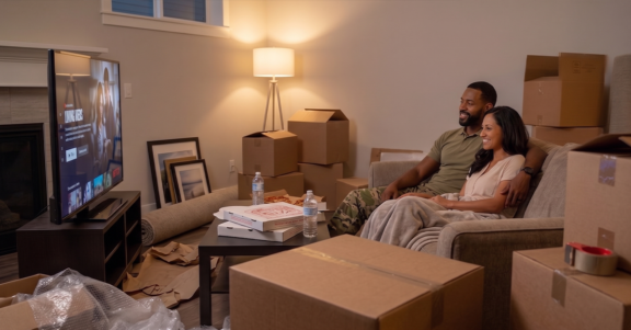 Settle into your new home with the hero savings and rewards you deserve. A military couple relaxes on a couch among moving boxes in their new living room, highlighting the hero savings achieved when choosing Homes for Heroes vs other programs.