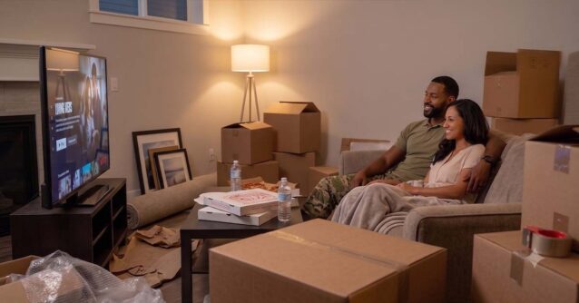 A military couple relaxes on a couch among moving boxes in their new living room, highlighting the hero savings achieved when choosing Homes for Heroes vs other programs.